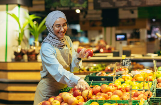 Female seller in hijab browsing and checking apples in supermarket, woman in apron smiling at work in store in fruit and vegetable department Female seller in hijab browsing and checking apples in supermarket, woman in apron smiling at work in store in fruit and vegetable department.