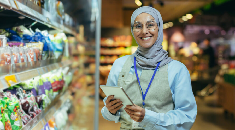A young Arab woman in a hijab works in a supermarket, salesperson, consultant, manager. Standing with a folder in his hands and a badge in the grocery department. He looks at the camera, smiles.
